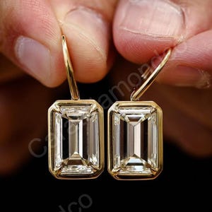 May include: A pair of gold-colored earrings with rectangular, clear gemstones. The gemstones are set in a gold bezel setting and have a step-cut design. The earrings are held against a dark background.