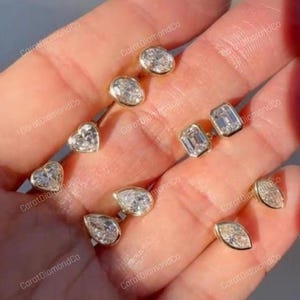 Puede incluir: Un conjunto de pendientes de oro con diamantes de diversas formas: ovalados, de corazón, talla esmeralda, pera y marquesa. Los pendientes se muestran en una mano. El texto "Carat DiamondCo" es visible.