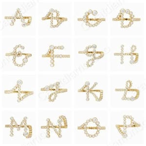 May include: A selection of gold-coloured rings, each featuring a different letter of the alphabet. The letters are crafted from small, round, clear gemstones. The rings are presented against a white backdrop.