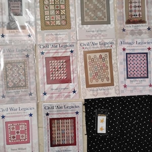 May include: A collection of Civil War Legacies quilt pattern booklets, featuring various quilt designs in red, blue, and beige. The booklets are arranged on a light-colored surface, with a black fabric with white polka dots and a small package of star-shaped buttons.
