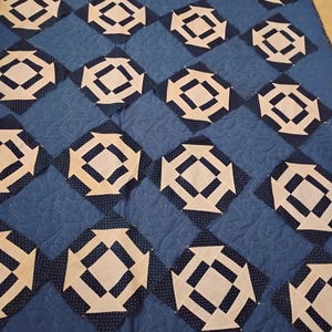 May include: A blue and white quilt with a geometric pattern. The quilt is made up of squares with a white center and a blue border. The squares are arranged in a repeating pattern.