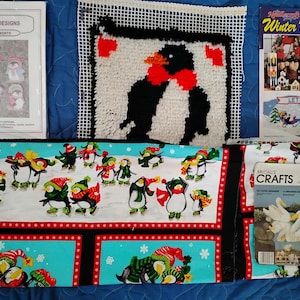 May include: An assortment of craft supplies featuring a penguin theme. Includes a Laura Doyle Designs pattern for penguin ornaments, a needlepoint piece, a "Winter Wonderland" kit, and fabric panels with penguin designs. The fabric has a red border and a turquoise background.