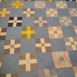 May include: A patchwork quilt with a light blue background and a grid pattern of squares. Each square features a cross design in various patterns and colors, including yellow, black, white, and floral prints. The quilt is a mix of textures and patterns.