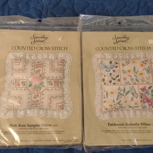 May include: Two cross-stitch pillow kits in plastic packaging. One is the Pink Rose Sampler Pillow, the other is the Patchwork Butterfly Pillow. Both kits feature floral designs and lace trim. The finished size is 12 x 12 inches.