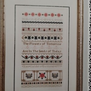 May include: A framed cross-stitch sampler with a white background and colorful embroidery. The sampler features a quote: "The Flowers of Tomorrow Are in The Seeds of Today." It also includes a decorative border with butterflies and a floral design. The sampler is signed "Man Caldera - Summer 1991."