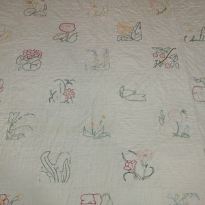May include: A white quilt featuring embroidered floral designs in various colors, including pink, red, green, and yellow. The quilt is divided into square blocks, each showcasing a different flower or plant. The stitching is visible, adding texture to the handmade textile.