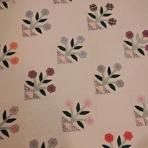 May include: A white quilt featuring a repeating pattern of flower baskets. Each basket contains three hexagonal flowers in various colors and patterns, including pink, blue, and orange, with dark green leaves and stems.
