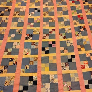 May include: A patchwork quilt with a grid pattern. The quilt features squares of various patterned fabrics in blue, yellow, and other colors, separated by pink and yellow strips. The quilt is likely handmade and has a vintage aesthetic.