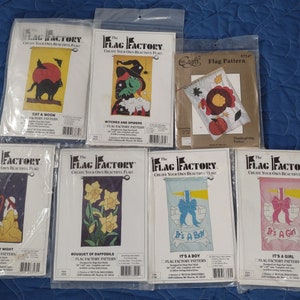 May include: A collection of packaged flag patterns from "The Flag Factory." Designs include a black cat with a moon, witches and spiders, a turkey, stars by night, daffodils, "It's a Boy," and "It's a Girl." Each pattern is in a plastic sleeve.