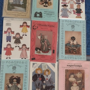 May include: A collection of vintage sewing patterns for dolls, featuring various designs and styles. The patterns include images of the finished dolls and are packaged in plastic sleeves. Titles include "Sweet Dreams", "Dara and Rene", and "Sugarlamps".