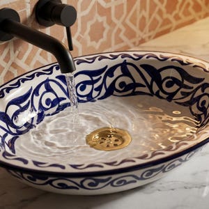 May include: Oval ceramic sink with a blue and white decorative pattern. Water flows from a black faucet into the sink, which has a gold-colored drain. The sink is set on a marble countertop, with a patterned tile wall in the background.