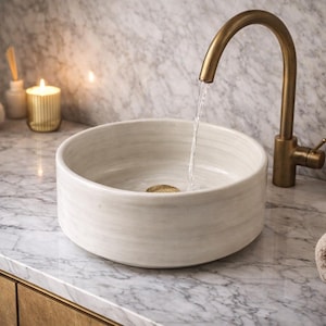 Puede incluir: Un lavabo de baño moderno con un grifo dorado. El lavabo de cerámica blanca es redondo y está sobre una encimera de mármol. También se ven una toalla enrollada, un dispensador de jabón y una vela.