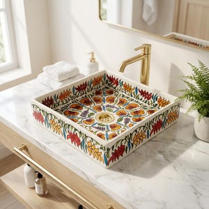 May include: A square ceramic bathroom sink featuring a floral design in red, yellow, orange, and blue. The sink is set into a white marble countertop with a gold tap. A gold drain and towel rail are also visible.
