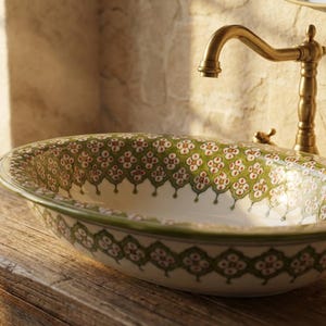 May include: Oval ceramic bathroom sink with a decorative green and red floral pattern. The sink is set on a wooden cabinet and features a brass faucet. The design is reminiscent of vintage or farmhouse styles.