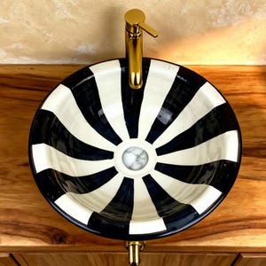 Puede incluir: Un lavabo de cerámica a rayas blancas y negras con un grifo dorado. Una toalla enrollada descansa sobre una bandeja dorada a la izquierda. Un plato pequeño con flores secas está a la derecha. El lavabo está sobre un mueble de madera.