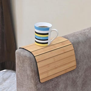Puede incluir: Una bandeja de brazo de sofá de bambú con una taza de té. La bandeja marrón claro está diseñada para colocarse en un reposabrazos de sofá. La taza blanca tiene rayas horizontales azules, amarillas y negras.