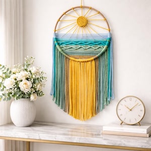 Könnte beinhalten: Ein handgefertigter Wandbehang mit einem Sonnenmotiv. Der kreisförmige Rahmen hat eine gelbe Sonne in der Mitte mit Strahlen. Darunter erzeugen Schichten aus blauem, grünem und gelbem Garn ein strukturiertes, fließendes Design. Es ist ein dekoratives Element für ein Zuhause.