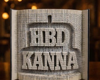 Puede incluir: Un libro abierto con las páginas plegadas para formar las palabras "HBD KANNA". Las letras en relieve destacan sobre las páginas de color beige claro. Un regalo artístico y original.