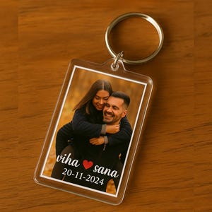 Könnte beinhalten: Ein klarer Acryl-Schlüsselanhänger mit einem silbernen Metallring. Der Schlüsselanhänger zeigt ein Foto eines lächelnden Paares. Der Text "viha ❤️ sana" und das Datum "20-11-2024" sind auf dem Foto aufgedruckt. Der Hintergrund ist eine verschwommene Szene im Freien.
