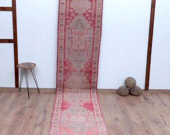 Puede incluir: Una alfombra de pasillo larga y estrecha con una paleta de colores rosa descolorido y beige apagado. El tejido muestra un patrón central de medallón geométrico con bordes intrincados, exhibiendo una textura desgastada en toda la superficie.