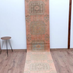 Op de afbeelding: Een lange, rechthoekige loper met een vervaagde, vintage uitstraling. De loper heeft een zalmkleurige achtergrond met herhaalde geometrische patronen in groen- en beigetinten. De loper wordt verticaal gepresenteerd, wat de lengte en het ontwerp benadrukt.