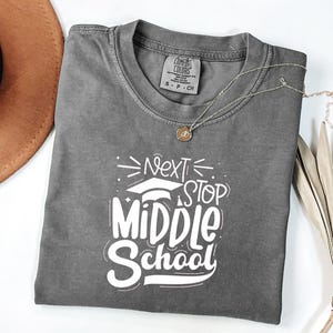 Op de afbeelding: Een grijs Comfort Colors T-shirt met de tekst "Next Stop Middle School" in het wit. Het T-shirt heeft een ronde hals en korte mouwen. Een gouden ketting met een hanger ligt over het T-shirt.