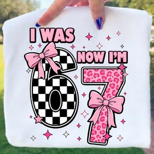 以下が含まれることがあります： 「I WAS NOW I'M」という文字がプリントされた白いTシャツ。数字の6は黒と白のチェック柄でピンクのリボンが付いており、数字の7はピンクのヒョウ柄でピンクのリボンが付いています。周囲には小さなピンクの星のアイコンが散りばめられています。