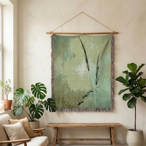 Puede incluir: Un tapiz de pared tejido con un diseño inspirado en la naturaleza. La obra de arte presenta un fondo abstracto verde y blanco con siluetas de plantas negras altas y delgadas. El tapiz está enmarcado por una barra de madera y tiene un borde con flecos.
