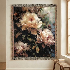 Puede incluir: Una manta tejida con un diseño floral que presenta grandes peonías color crema sobre un fondo oscuro. La manta tiene un borde con flecos con una mezcla de colores y está colgada en una pared.