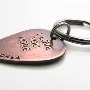 May include: A copper-colored guitar pick-shaped keychain with the words "I LOVE YOU MORE" and the name "ANDREA" engraved on it. The keychain has a silver-colored split ring.