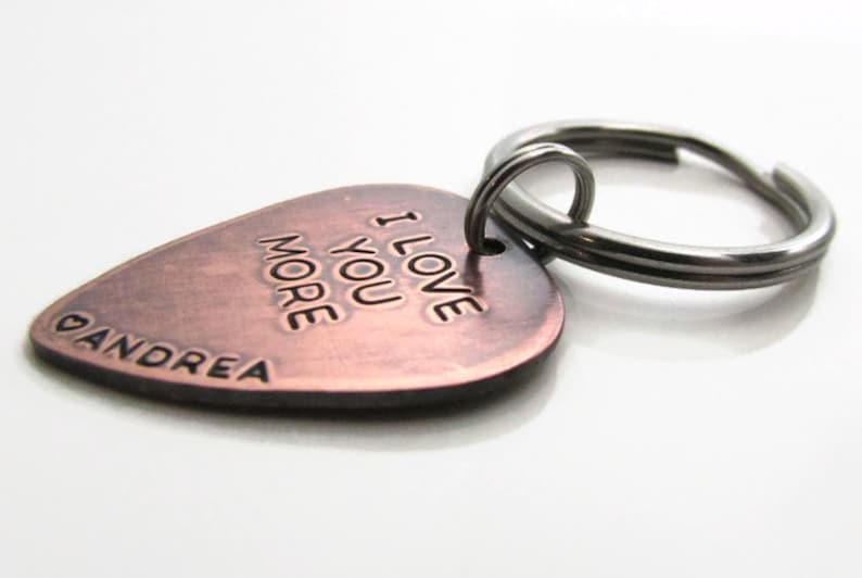 May include: A copper-colored guitar pick-shaped keychain with the words "I LOVE YOU MORE" and the name "ANDREA" engraved on it. The keychain has a silver-colored split ring.