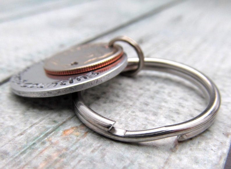 May include: A silver key chain with a round, engraved metal charm and a copper-colored coin attached. The charm has a smooth, polished surface.