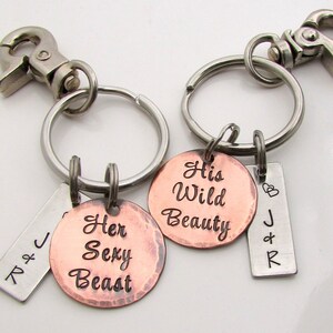 May include: Two keychains with silver metal clasps and rings. One keychain has a copper-colored round charm with the words "Her Sexy Beast". The other keychain has a copper-colored round charm with the words "His Wild Beauty". Both keychains have a small silver tag with initials.