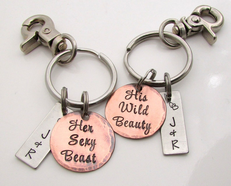 May include: Two keychains with silver metal clasps and rings. One keychain has a copper-colored round charm with the words "Her Sexy Beast". The other keychain has a copper-colored round charm with the words "His Wild Beauty". Both keychains have a small silver tag with initials.