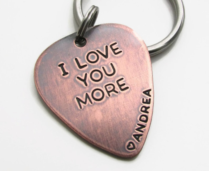 May include: A copper-colored guitar pick keychain with the words "I LOVE YOU MORE" engraved on it. The name "ANDREA" is also engraved on the pick. The keychain has a silver-colored ring.