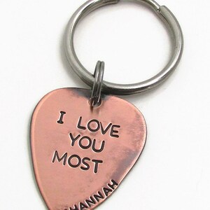 May include: A copper-colored heart-shaped keychain with the words "I LOVE YOU MOST" engraved on it. The name "Hannah" is inscribed below, with a small heart symbol. The keychain has a silver-colored ring.