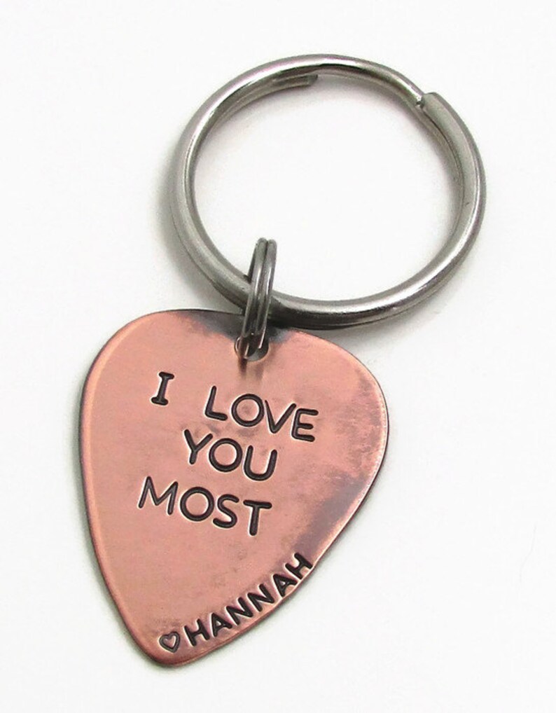 May include: A copper-colored heart-shaped keychain with the words "I LOVE YOU MOST" engraved on it. The name "Hannah" is inscribed below, with a small heart symbol. The keychain has a silver-colored ring.