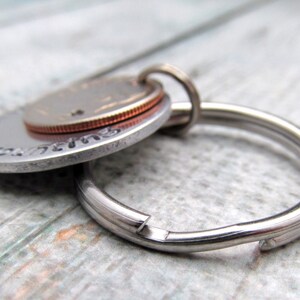 May include: A silver key chain with a round, silver charm featuring a copper-colored coin. The charm has a decorative border and the coin is visible in the center.
