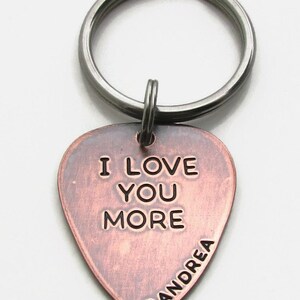 May include: A copper-colored guitar pick keychain with the words "I LOVE YOU MORE" engraved in black. The name "Andrea" is also engraved. The keychain has a silver-colored ring.
