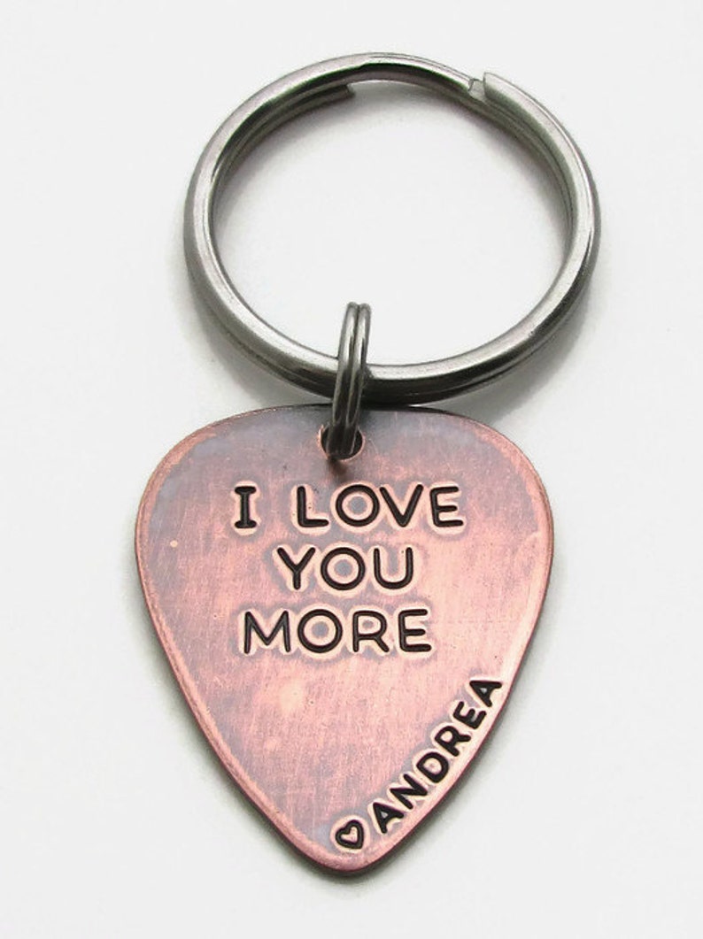 May include: A copper-colored guitar pick keychain with the words "I LOVE YOU MORE" engraved in black. The name "Andrea" is also engraved. The keychain has a silver-colored ring.