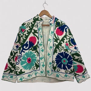 Könnte beinhalten: Weiße Jacke mit farbenfroher Blumenstickerei. Die Jacke hat einen Stehkragen und lange Ärmel. Die Stickerei umfasst blaue, rosa, grüne und türkisfarbene Blumen und Blätter. Die Jacke hängt an einem Holzbügel.