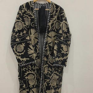 May include: A black embroidered coat with a floral design in a gold color. The coat has a striped lining and is hanging on a wooden hanger. The embroidery covers the entire coat, creating a luxurious look.