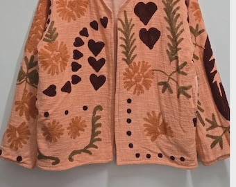 May include: A peach-colored, open-front jacket with embroidered floral and heart designs. The jacket features a V-neck and long sleeves. The embroidery includes shades of brown and orange, creating a textured, bohemian aesthetic. The jacket is hanging on a wooden hanger.