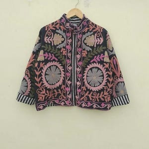 Könnte beinhalten: Schwarze Jacke mit aufwendiger Blumenstickerei in Rosa, Grau und Grün. Die Jacke hat einen hohen Kragen und gestreifte Akzente an den Manschetten und der Vorderseite. Die Stickerei umfasst große kreisförmige Blumendesigns.