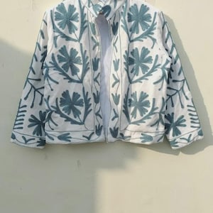 Könnte beinhalten: Weiße Jacke mit tealfarbener Blumenstickerei. Die Jacke hat einen Kragen und lange Ärmel. Das florale Design wiederholt sich auf der Vorderseite, dem Rücken und den Ärmeln. Die Jacke hängt an einem Holzbügel.