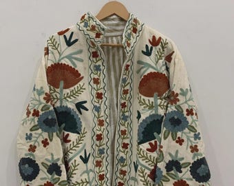 May include: Cream-colored jacket with intricate floral embroidery in shades of blue, red, and green. The jacket features a stand-up collar and is open in the front, revealing a striped lining. The embroidery is detailed and covers the front, sleeves, and collar.