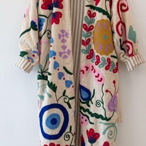 May include: A long, cream-colored coat featuring vibrant, multi-colored floral and abstract embroidery. The garment has long sleeves with striped cuffs, a stand-up collar, and an open front, revealing a vertical striped lining on the interior.