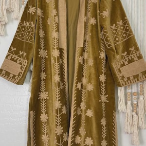 Könnte beinhalten: Olivgrüner Samt-Duster-Coat mit floraler Stickerei. Der lange Mantel hat einen Reverskragen, lange Ärmel und eine kontrastierende beigefarbene Paspel. Die Stickerei ist hellbeige.