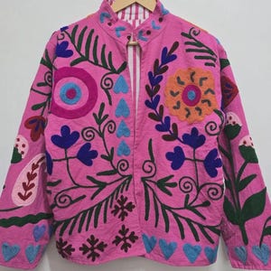 Könnte beinhalten: Eine leuchtend pinkfarbene Jacke mit aufwendiger Blumenstickerei in Blau-, Grün-, Orange- und Rosatönen. Die Jacke hat einen Stehkragen und einen Knopfverschluss. Das Design umfasst gestickte Blumen, Blätter, Herzen und andere dekorative Elemente.