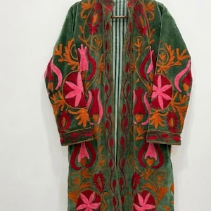 Könnte beinhalten: Ein langärmeliger, vorne offener Mantel aus olivgrünem Samt mit lebhaften Stickereien in Rosa-, Rot- und Orangetönen. Die floralen und botanischen Muster bedecken die Vorderteile und Ärmel, während das Innenfutter einen weiß-grün gestreiften Stoff zeigt.